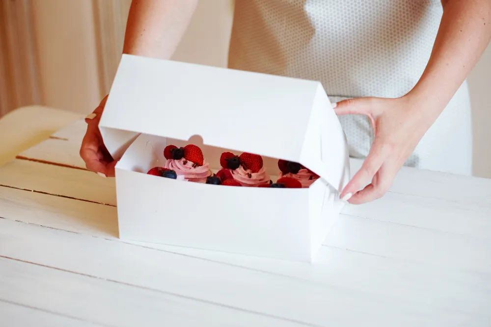 Osoba otwiera wieko białego pudełka na muffiny, z różowymi muffinkami wewnątrz.